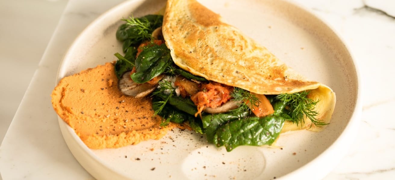 Photo from the kitchen, showing a plate standing on the counter. In the centre of the plate is a half-folded omelette inside which vegetables, cheese and herbs are wrapped. Next to the omelette on the plate is toast slightly covered by the omelette.